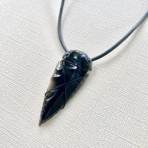 Puede incluir: Un colgante de punta de flecha de obsidiana negra en un collar de cordón de cuero negro. La punta de flecha está envuelta con alambre negro.