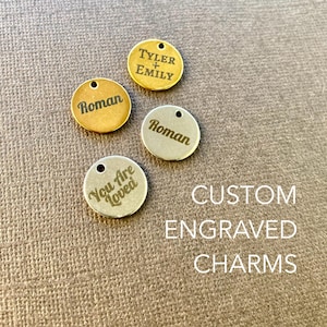Puede incluir: Cuatro abalorios de metal grabados personalizados. Los abalorios son redondos y tienen diferentes diseños. Un abalorio dice "Tyler + Emily", otro dice "Roman", otro dice "Roman" y el último dice "You Are Loved". Los abalorios están sobre una superficie marrón texturizada. CUSTOM ENGRAVED CHARMS está escrito en blanco debajo de los abalorios.