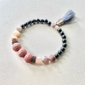May include: A beaded bracelet with a gray tassel. The bracelet features black hematite beads, pink lava rock beads, and white and pink opal beads.