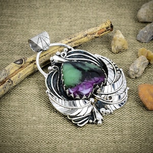 Puede incluir: Un colgante de plata con una piedra central en forma de corazón de color verde vibrante y morado intenso. La piedra está engastada en una montura de plata pulida decorada con intrincados grabados de hojas y pequeñas esferas de plata.