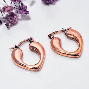 Puede incluir: Un par de pendientes de aro en forma de corazón de color cobre. Los pendientes presentan un acabado metálico liso con un mecanismo de cierre de bisagra en la parte superior. El diseño es sencillo y curvado, con un tono marrón rojizo.