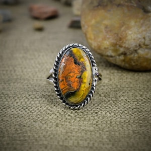 May include: An oval-shaped ring featuring a polished bumblebee jasper stone with swirling patterns of vibrant orange, yellow, and black. The stone is set in a silver-toned metal band with a decorative twisted rope border and small bead accents.