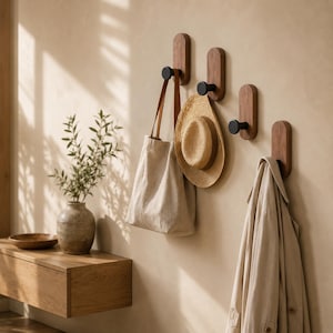 Puede incluir: Cuatro percheros de pared con placas de madera ovaladas verticales de bordes redondeados y ganchos cilíndricos de color negro mate. Los percheros están alineados y sostienen un bolso de tela beige, un sombrero de paja y una gabardina beige.