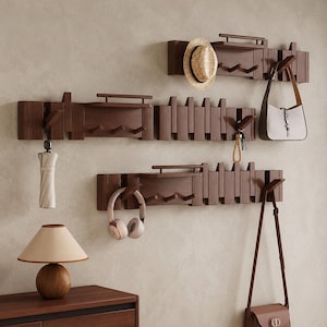 May include: Three dark brown wooden wall-mounted coat racks with horizontal slats, small shelves, and multiple pegs. The racks hold a straw hat, a beige umbrella, a set of keys, a white handbag, pink headphones, and a brown leather shoulder bag.