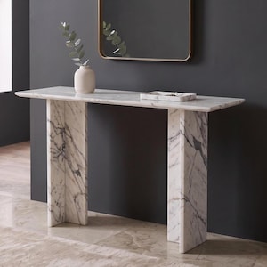 May include: A white marble console table with grey veining, featuring a rectangular top supported by two vertical slab legs. On the surface sits a matte white ceramic vase containing a green eucalyptus sprig and a small rectangular marble tray.