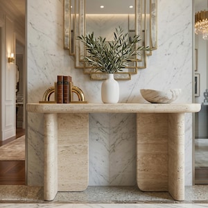 May include: A beige travertine console table with a smooth top and two curved legs. On the table are two gold arch bookends holding two brown books, a white ceramic vase with green olive branches, and a beige marble bowl with a veined pattern.