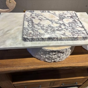 May include: A rectangular marble tray featuring a white and grey brecciated stone pattern with a fluted edge, resting on a smooth, flat, light-colored marble slab. The tray displays a high-contrast, multi-tonal stone surface with distinct veining.
