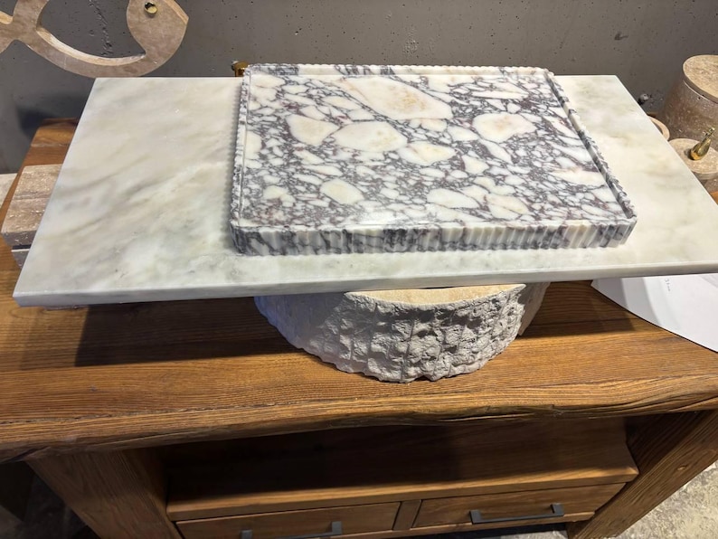 May include: A rectangular marble tray featuring a white and grey brecciated stone pattern with a fluted edge, resting on a smooth, flat, light-colored marble slab. The tray displays a high-contrast, multi-tonal stone surface with distinct veining.