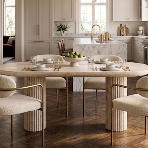 May include: An oval dining table with a light beige stone top and two fluted cylindrical bases, surrounded by four cream-colored textured chairs with gold metal frames. The table is set with white plates, bowls, glasses, and gold-toned cutlery.