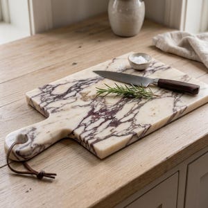 May include: A rectangular marble serving board with a cream and deep purple veined pattern, featuring a handle with a brown leather cord. A stainless steel knife with a dark wood handle and a sprig of fresh green rosemary rest on top.