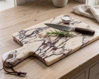 May include: A rectangular marble serving board with a cream and deep purple veined pattern, featuring a handle with a brown leather cord. A stainless steel knife with a dark wood handle and a sprig of fresh green rosemary rest on top.