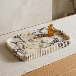 May include: A rectangular marble tray featuring a white base with deep purple veining and a fluted, ribbed edge. Resting on the tray is a small, square amber glass bottle and a delicate gold-toned chain necklace with a thin, simple link design.
