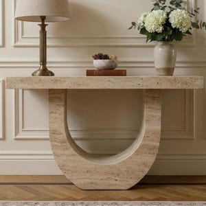 May include: A beige travertine console table with a U-shaped base. On the table sits a brass lamp with a beige shade, a small bowl containing pinecones, a brown book, and a ceramic vase filled with white hydrangea flowers and green leaves.