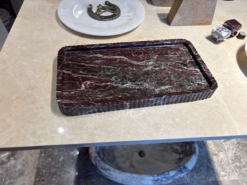 May include: A rectangular tray crafted from deep red marble with prominent white and dark grey veining. The tray features a distinctively fluted or ribbed edge design that runs along the entire perimeter, providing a textured border to the smooth surface.