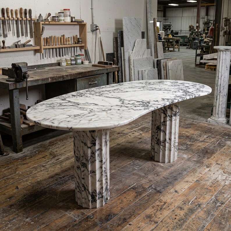 May include: An oval white marble table with prominent grey veining, supported by two fluted cylindrical marble pedestals. The tabletop features a smooth, polished surface, while the base columns exhibit a vertical grooved texture throughout.