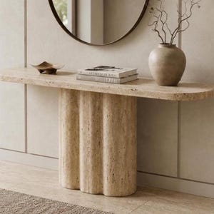 May include: A beige travertine console table with a smooth, elongated oval top and a thick, vertically fluted base. The table surface features natural porous textures and holds a small decorative bowl, two stacked books, and a ceramic vase.
