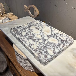 May include: A rectangular tray featuring a white and dark purple marble terrazzo pattern with a fluted edge. The tray is positioned on a smooth white marble slab, showcasing a high-contrast stone texture with irregular white fragments embedded in a dark base.