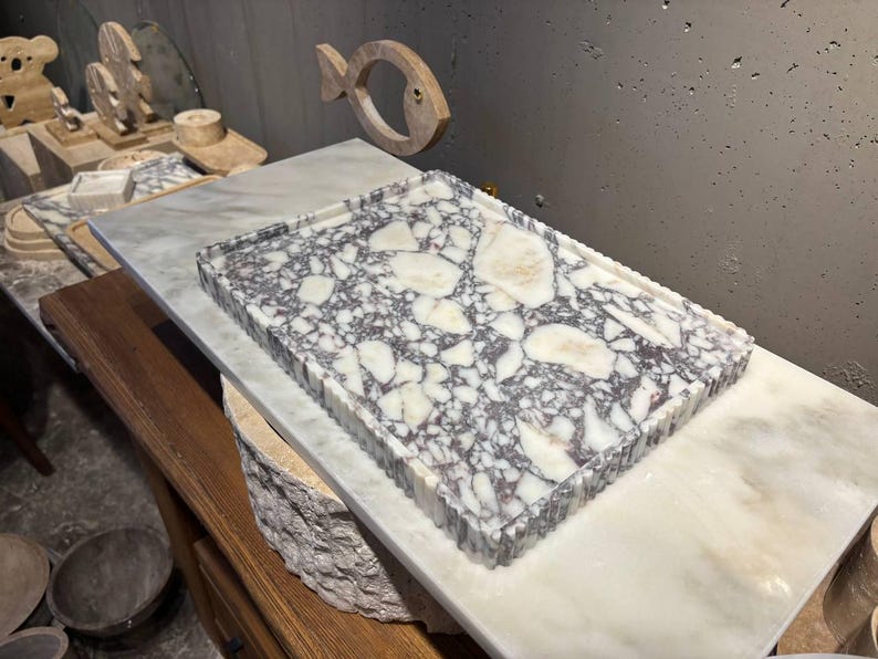 May include: A rectangular tray featuring a white and dark purple marble terrazzo pattern with a fluted edge. The tray is positioned on a smooth white marble slab, showcasing a high-contrast stone texture with irregular white fragments embedded in a dark base.
