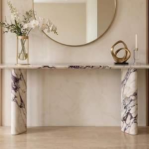 May include: A long console table featuring a white marble top with purple veining and two thick, cylindrical marble legs with matching purple veining. The table surface holds a glass vase with white flowers, a gold abstract sculpture, and a candle.