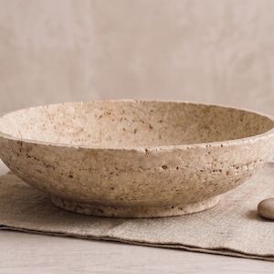 May include: A round, shallow bowl crafted from beige, porous stone with a textured, pitted surface. The bowl features a wide, open interior and a sturdy, raised circular base, showcasing natural variations in the stone's color and grain.