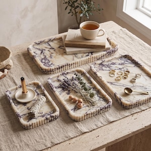 May include: Three rectangular marble trays with purple veining and fluted edges. One tray holds a ceramic incense holder with a stick and a sage bundle. Another tray contains dried lavender and wooden beads. The third tray holds gold rings and a spoon.