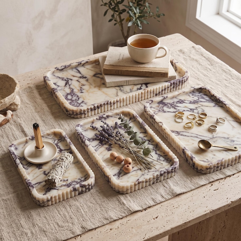 May include: Three rectangular marble trays with purple veining and fluted edges. One tray holds a ceramic incense holder with a stick and a sage bundle. Another tray contains dried lavender and wooden beads. The third tray holds gold rings and a spoon.