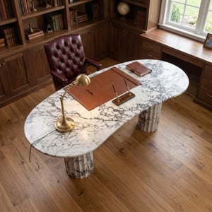 May include: An oval desk with a white and grey marble top supported by two fluted marble columns. The desk features a brown leather desk pad, a brass desk lamp, a double pen stand with two pens, and a closed brown leather notebook on the surface.