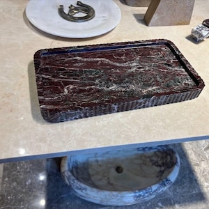 May include: A rectangular tray crafted from deep red and dark green marble with prominent white veining. The tray features a raised, fluted edge design that runs along the entire perimeter, providing a textured border to the polished surface.