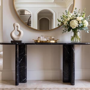 May include: A black marble console table with two curved pillar legs, topped with a beige abstract sculpture, a gold tray holding clear crystals and a glass bowl, and a clear glass vase filled with white flowers and eucalyptus.
