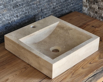 Könnte beinhalten: Ein quadratisches Waschbecken aus beigem Stein mit einem glatten, geneigten Becken, einem zentralen Abflussloch und einer kleineren kreisförmigen Öffnung für eine Armatur. Das Waschbecken hat einen flachen, breiten Rand und eine natürliche Oberfläche.
