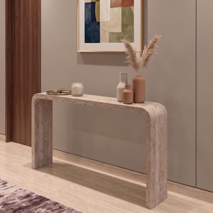 May include: A beige travertine console table with a curved waterfall design. On the surface sits a small gold tray with keys, a white candle in a glass jar, and three ceramic vases in varying shades of beige and terracotta with dried grass.