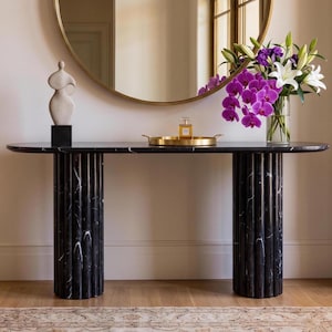 May include: A black marble console table with two fluted cylindrical legs, topped with a beige abstract sculpture on a black base, a gold tray holding a perfume bottle, and a glass vase filled with purple orchids and white lilies.