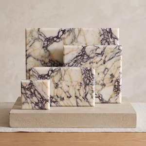 May include: A collection of four rectangular and square marble slabs featuring a polished white surface with prominent, intricate veining in shades of deep purple and golden brown, arranged in a tiered formation on a thick, light beige base.
