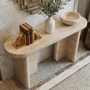May include: A beige travertine console table with a rounded top and two curved legs. On the table are two brown books held by gold metal bookends, a white ceramic vase with green foliage, and a beige stone bowl with an irregular rim.