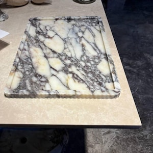 May include: A rectangular marble serving tray featuring a polished surface with a prominent white base color and intricate, dark grey to black veining throughout. The tray has a raised, fluted edge border that frames the central flat area.