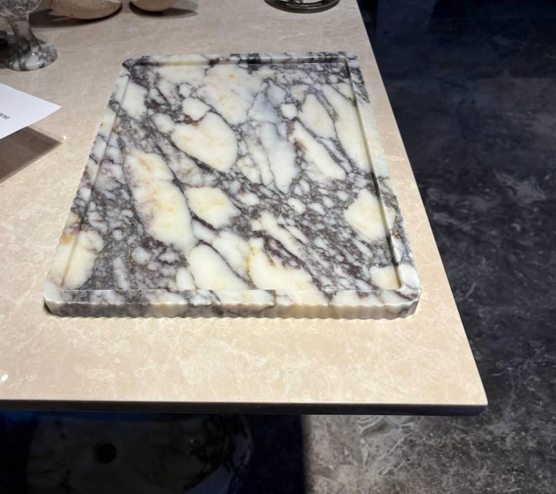 May include: A rectangular marble serving tray featuring a polished surface with a prominent white base color and intricate, dark grey to black veining throughout. The tray has a raised, fluted edge border that frames the central flat area.