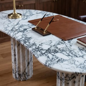 May include: An oval desk with a white and black marble top and two fluted marble pedestal bases. On the desk sits a brown leather desk pad, a wooden pen holder with two gold and black pens, a gold lamp base, and a brown leather-bound notebook.