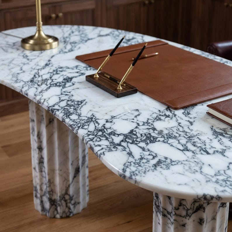May include: An oval desk with a white and black marble top and two fluted marble pedestal bases. On the desk sits a brown leather desk pad, a wooden pen holder with two gold and black pens, a gold lamp base, and a brown leather-bound notebook.