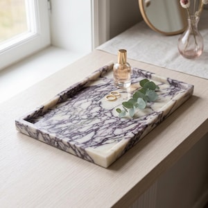 May include: A rectangular tray crafted from white marble with prominent deep purple veining. The tray features raised edges and a smooth, polished surface. Resting on the tray are a small glass perfume bottle with a gold cap and two gold rings.