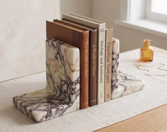 Könnte beinhalten: Zwei L-förmige Buchstützen aus weißem Marmor mit ausgeprägter dunkelvioletter Maserung. Die Buchstützen halten vier gebundene Bücher mit den Titeln The Marble Garden, A Study in Veins, Geology and Aesthetics und Timeless Textures.