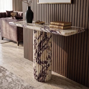 May include: A console table featuring a white and purple marble top and a matching fluted cylindrical base. The table surface holds a black vase with thin branches, a small gold-toned dish, and two stacked books with gold-embossed spines.