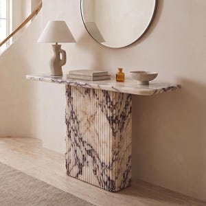 May include: A rectangular console table featuring a fluted base and a flat top, both crafted from white marble with prominent purple veining. The table stands on a floor, supporting a lamp, two stacked books, a small amber bottle, and a bowl.
