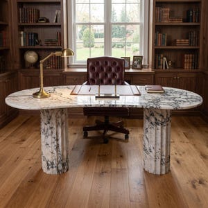May include: A large oval desk with a white and grey marble top supported by two fluted marble columns. The desk features a leather desk pad, a metal pen stand with two pens, a small notebook, and a brass desk lamp with a rounded shade.