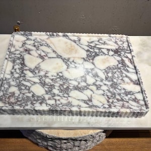 May include: A rectangular marble tray featuring a polished surface with a distinct brecciated pattern of white and deep purple-grey veining. The tray has a raised, fluted edge that provides a textured border around the smooth, flat interior surface.