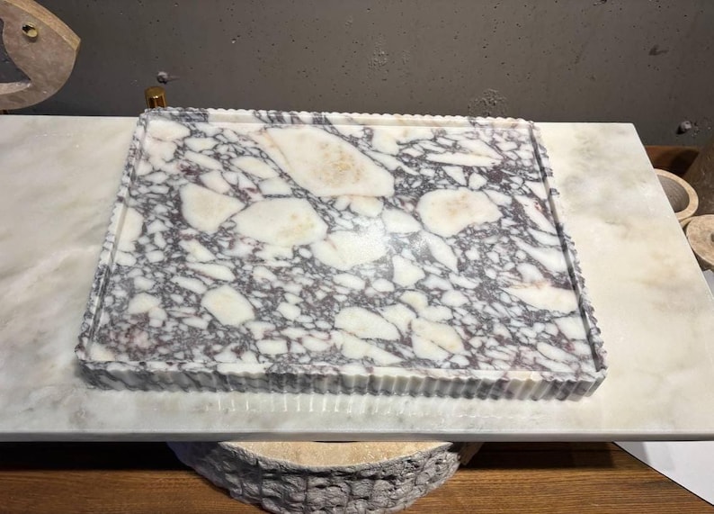 May include: A rectangular marble tray featuring a polished surface with a distinct brecciated pattern of white and deep purple-grey veining. The tray has a raised, fluted edge that provides a textured border around the smooth, flat interior surface.