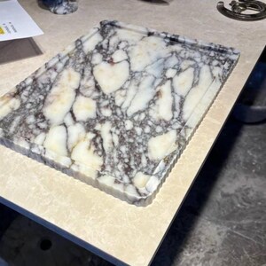 May include: A rectangular marble tray featuring a white base with prominent dark grey and black veining throughout. The tray has a raised, fluted edge design around the perimeter, providing a textured border to the smooth, polished stone surface.
