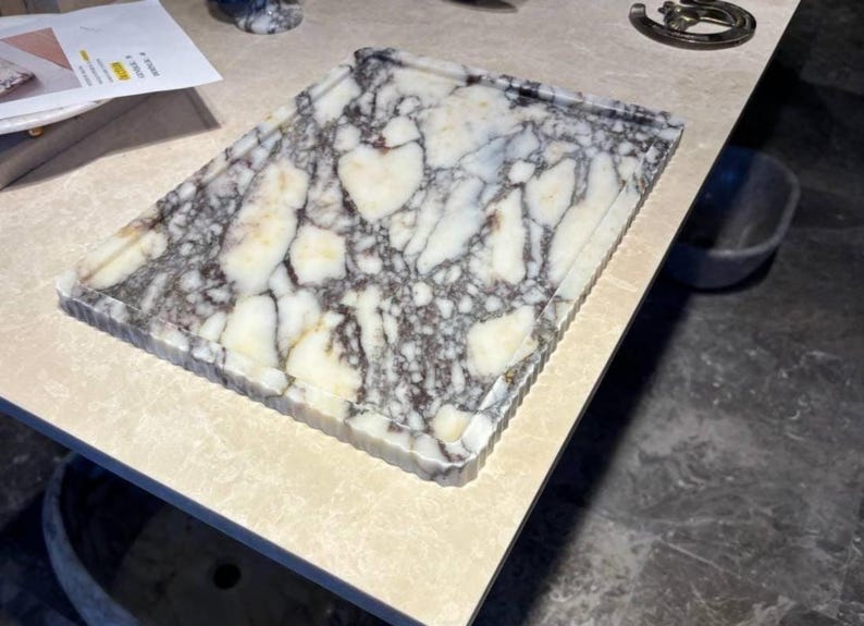 May include: A rectangular marble tray featuring a white base with prominent dark grey and black veining throughout. The tray has a raised, fluted edge design around the perimeter, providing a textured border to the smooth, polished stone surface.