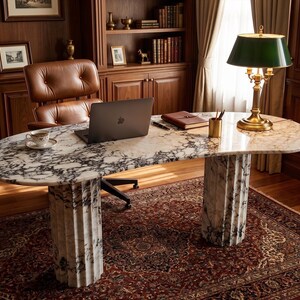 May include: An oval-shaped desk featuring a white and black veined marble top supported by two fluted marble columns. The desk surface holds a dark gray laptop, a small white teacup on a saucer, a brown leather notebook, and a gold pen holder.