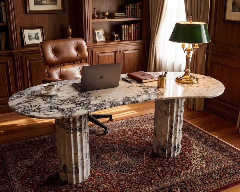 May include: An oval-shaped desk featuring a white and black veined marble top supported by two fluted marble columns. The desk surface holds a dark gray laptop, a small white teacup on a saucer, a brown leather notebook, and a gold pen holder.