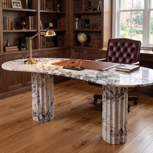 May include: An oval desk featuring a white and grey marble top supported by two fluted marble columns. The desk surface includes a brown leather desk pad, a gold-toned desk lamp, a pen stand with two pens, and a closed notebook.
